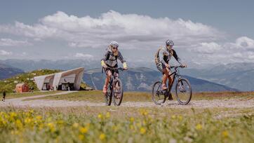 Zwei Fahrradfahrer unterwegs auf dem Kronplatz, hinter ihnen das Messner Mountain Museum